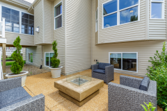 Outdoor courtyard with beige siding, large windows, woven patio seating, fire pit table, and landscaped greenery.