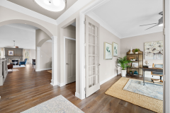 “Bright interior with hardwood floors, white walls, arched doorway, small office area with desk, rug, shelving, artwork, and potted plant.