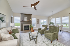 Living room with fireplace and dark wood mantel, green armchairs, beige sofa, glass coffee table, ceiling fan, chandelier over dining area, large windows with blinds, and wood‑look flooring.