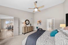 “Bedroom with beige walls, ceiling fan, large bed with patterned bedding and blue accent pillows, dresser, and doorway to adjoining room.