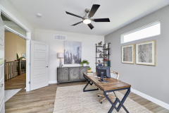 Home office with wooden desk, chair, computer monitor, framed artwork, ceiling fan with lights, large window, and wood flooring.